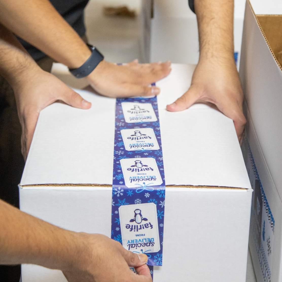 Two fulfillment team members working together to seal a white shipping carton with custom designed eco-friendly packing tape imprinted with a repeat pattern of gift tag with the words Special Delivery from fairlife (logo) and icons of snow flakes 