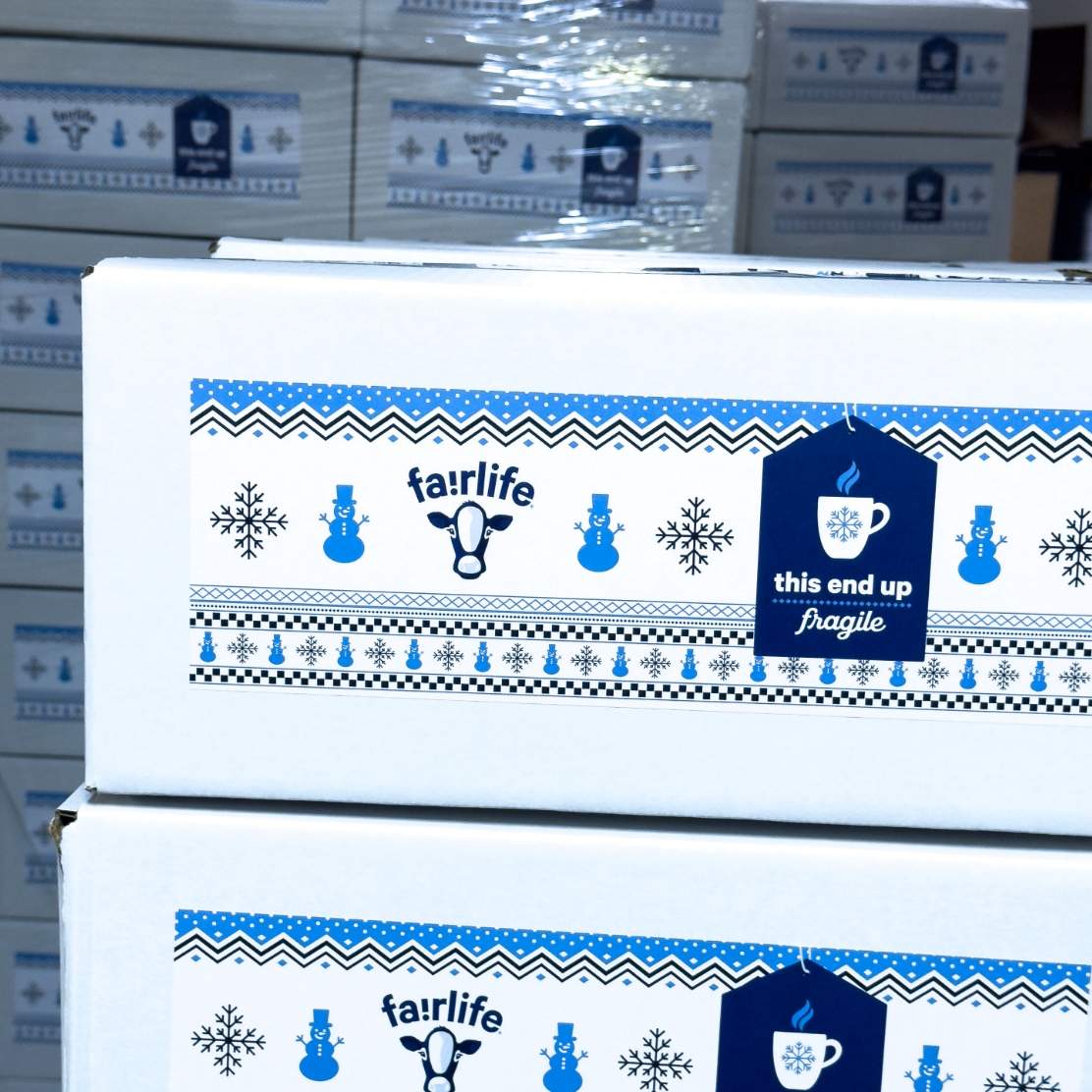 Closeup of two white shipping boxes with a holiday inspired mailing label imprinted with the fairlife cow logo, smowman and snowflake patterns and a gift tag image with a coffee cup and the words This End Up with a plastic wrapped pallet of stacked mailing boxes.