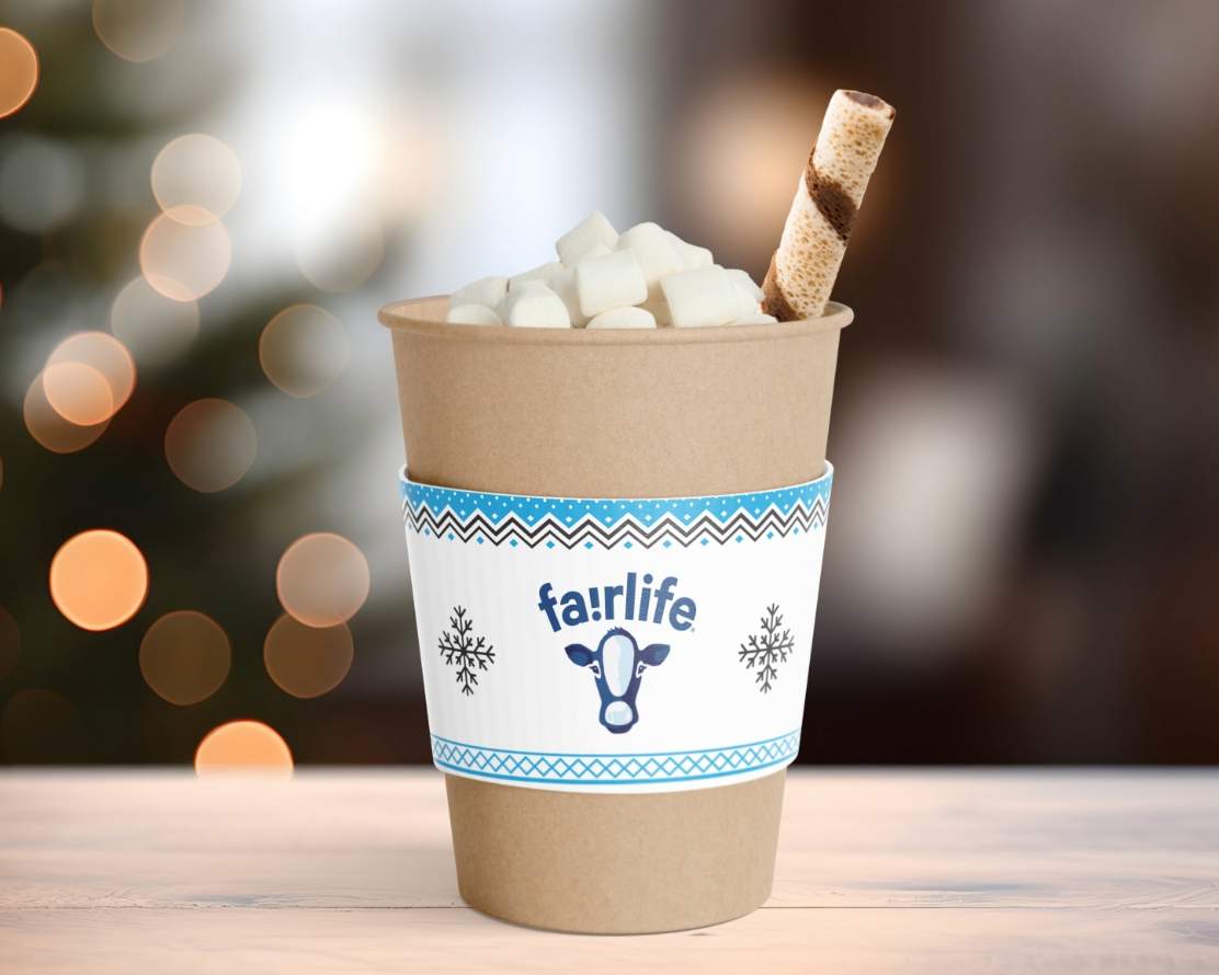 Kraft paper coffee cup with a custom designed fairlife cup sleeve filled with hot chocolate, marshmallows and a rolled wafrer cookie on a wooden counter with a blurred holiday tree and lights in the background