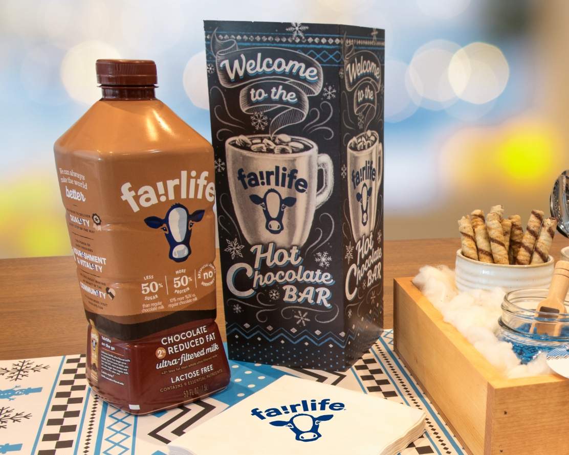 fairlife holiday promotional kit showing a close up of the assembled table tent, custom imprinted napkins, bottle of fairlife chocolate milk, table runner and corner of the wooden fairlife hot chocolate bar with holiday lights in the background