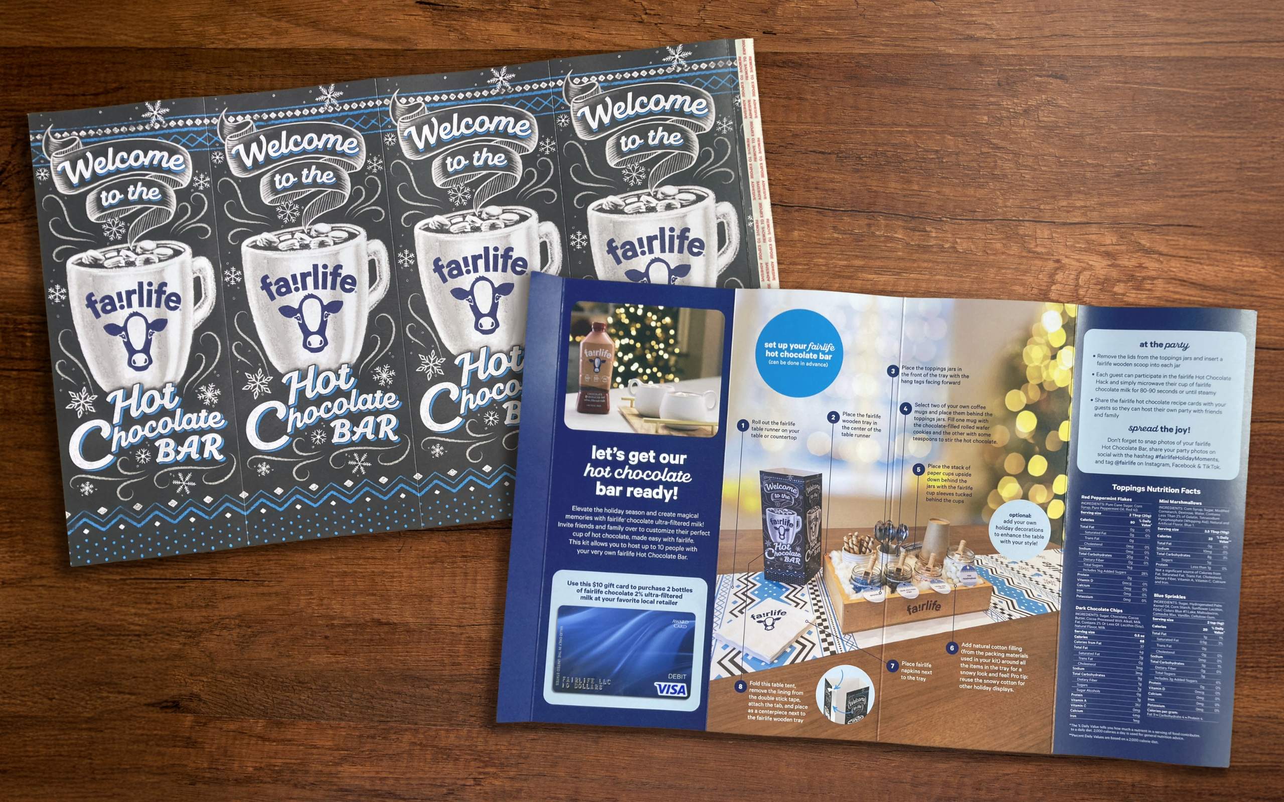 Printed samples on top of a wooden table top of 4-sided table tent showing a decorative chalkboard style illustration of a steaming cup of hot chocolate, the words Welcome to the fairlife hot chocolate bar and a large photo of the fairlife hot chocolate bar kit with set up instructions, gift card and nutritional facts