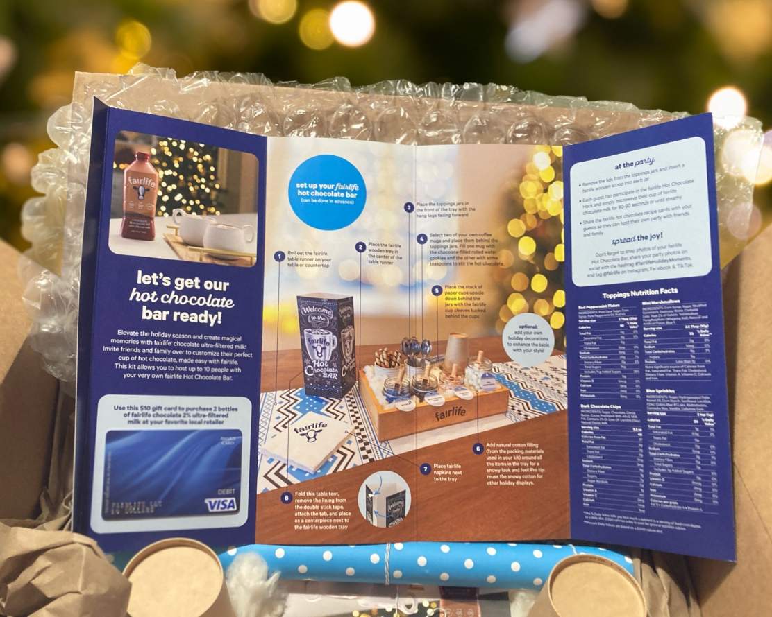 Open mailing box with an instructional brochure on top of the box contents. The brochure has a large photo of the fairlife hot chocolate bar kit with set up instructions on top.